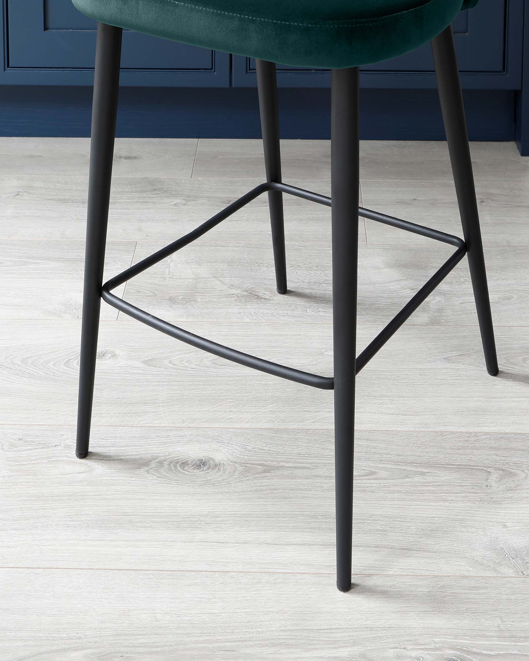 Close-up view of a contemporary bar stool with sleek metal legs in a matte black finish, supporting a round, plush seat upholstered in luxurious teal fabric. The geometric stability bars add both style and function to the design. The stool is positioned on a light wood laminate flooring, against a backdrop of dark blue cabinetry suggesting a modern and sophisticated kitchen or bar setting.