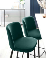 Two modern emerald green velvet dining chairs with black metal legs, in front of a sleek white table and a minimalist-style console table with a glass top and black metal frame. The console table is accessorized with a vase of white flowers, framed picture, vases, and books.