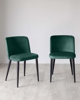 Two modern green velvet dining chairs with black metal legs, placed on a light wooden floor against a plain white wall.