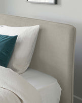 Elegant upholstered bed with a high, curved headboard in a light beige fabric. The bed is dressed with crisp white bedding complemented by a soft, textured white throw and a single teal accent pillow. A minimalist framed artwork is partially visible above the headboard.