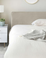 A minimalist bedroom featuring a neutral-toned upholstered bed headboard and a white bedside table with ribbed drawers and tapered wooden legs. The scene is complemented by a white bedspread, a round wall mirror with a thin frame, a white lamp with a cylindrical shade on the bedside table, and a small potted plant in a light blue ceramic pot.