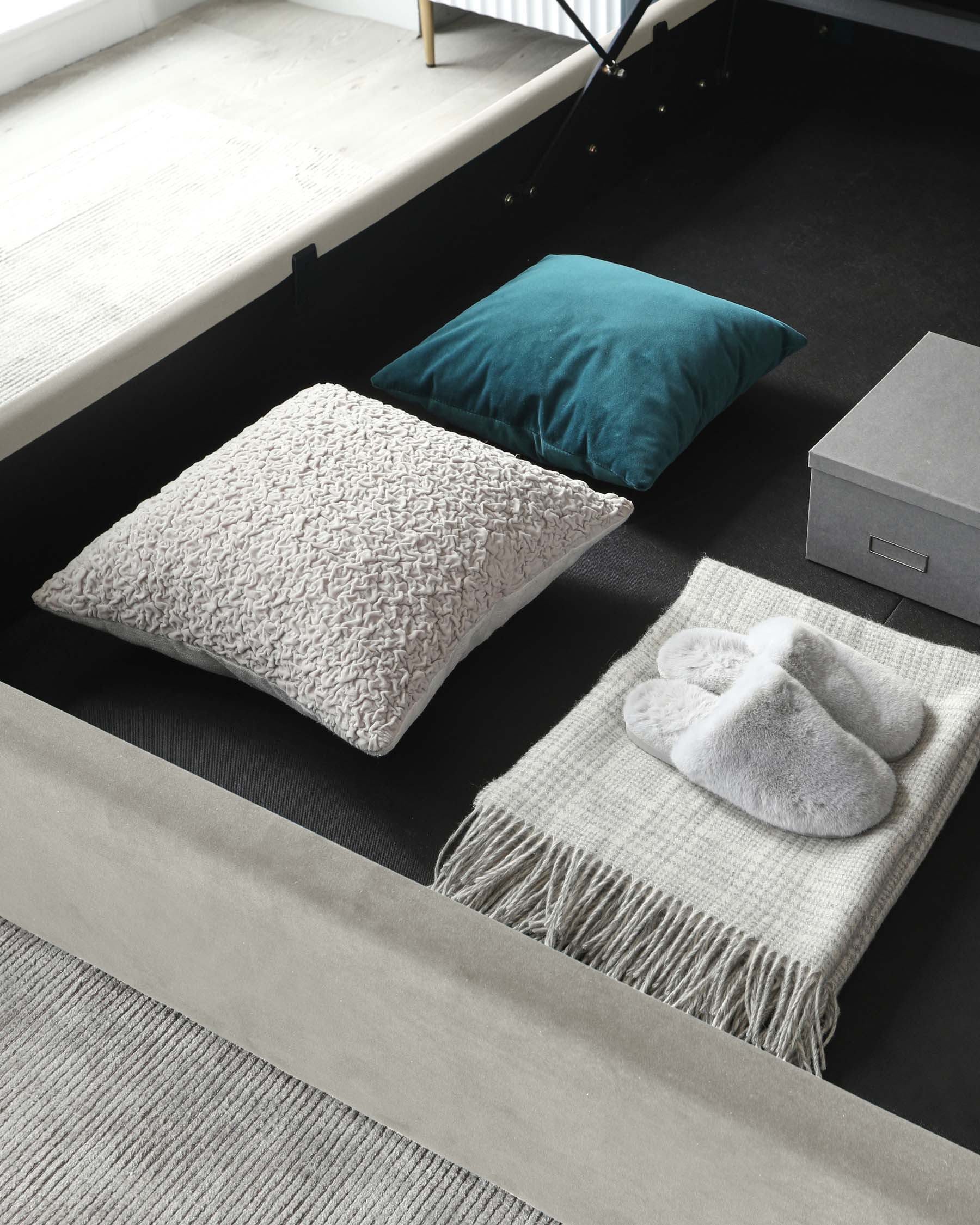 A minimalist low-profile platform bed with a light wooden base and black supporting structure. A dark fabric storage box is visible at the foot of the bed. The bedding is not visible, suggesting the focus is on the frame itself. The room has a simple, modern aesthetic with a plush grey carpet on the floor.