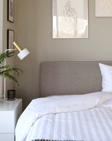 Contemporary bedroom featuring a stylish medium-grey upholstered headboard, paired with a white and grey striped bedding set on a bed frame that is not visible. Beside the bed, there is a sleek white bedside table with a reflective surface and geometric patterned front, topped with a cylindrical black vase.