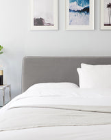 Modern minimalist bedroom featuring a queen-sized platform bed with a grey upholstered headboard, crisp white bedding, and a textured throw blanket. A clear glass side table with a gold base and a chic, modern lamp adorn one side, complementing the serene and stylish decor.