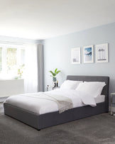 Modern minimalist style bedroom featuring a queen-sized bed with a sleek grey upholstered headboard and bed frame, dressed with crisp white bedding and accentuated with a light beige throw blanket. Two mirrored nightstands flank the bed, each bearing a decorative table lamp and minimalistic accessories.