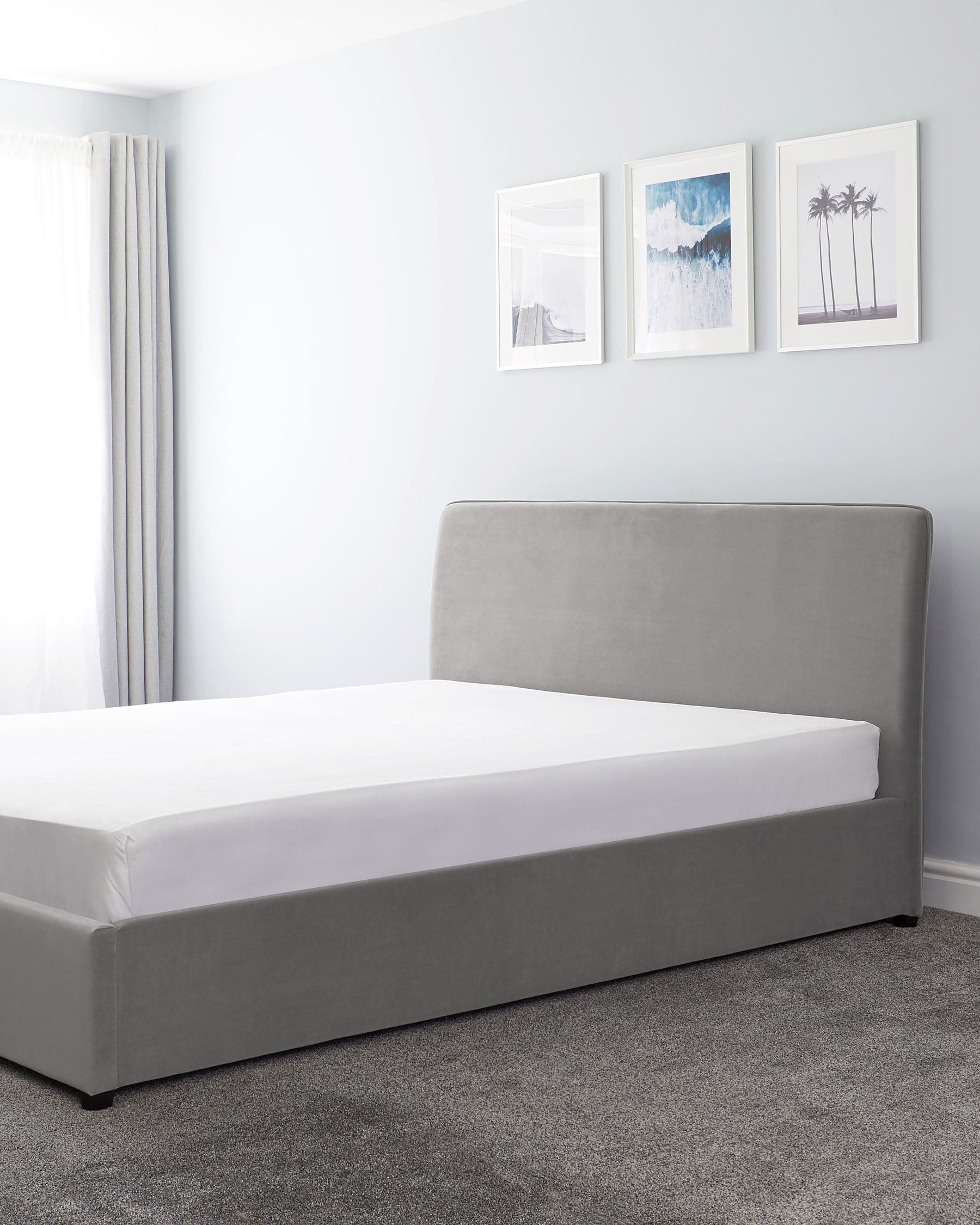 A modern, minimalist upholstered platform bed with a wide, slightly curved headboard and clean lines, displayed in a tidy bedroom with a neutral colour palette. The bed is dressed with a plain white fitted sheet and stands on a textured grey carpet.