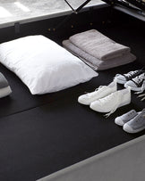 A modern storage bed with a dark grey upholstery and a hydraulic lifting mechanism revealing a hidden storage space underneath. The bed is accessorized with folded grey towels and a white pillow, accompanied by a pair of white and grey sneakers placed on the floor beside the bed.