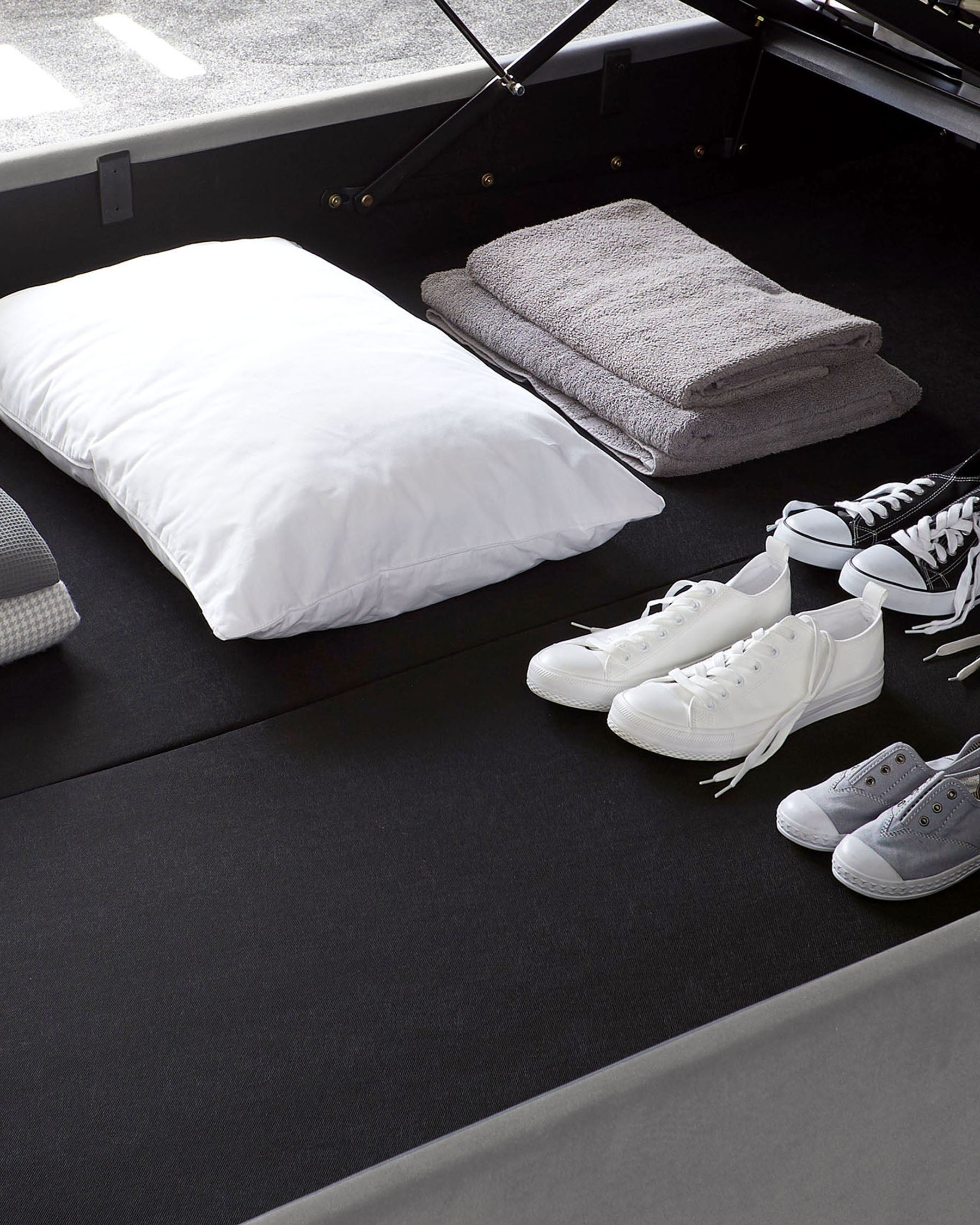 A modern storage bed with a dark grey upholstery and a hydraulic lifting mechanism revealing a hidden storage space underneath. The bed is accessorized with folded grey towels and a white pillow, accompanied by a pair of white and grey sneakers placed on the floor beside the bed.