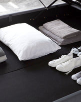 Black modern storage bed with a lifted frame revealing spacious storage underneath, accompanied by a white pillow and folded grey towels placed on top.