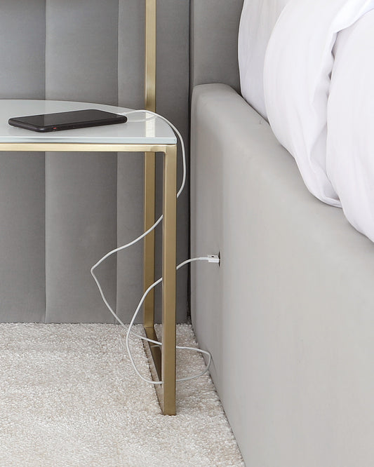 Elegant modern bedside table with a clear glass top and minimalist gold-tone metal frame, alongside a plush grey upholstered bed with crisp white bedding.