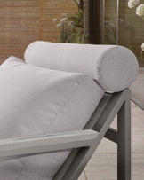 A close-up of a modern outdoor lounge chair featuring a solid dark grey aluminium frame and light grey cushions. The armrests are broad and flat, and there's a cylindrical bolster pillow atop the chair's angled backrest. The chair's sleek design and materials suggest both comfort and durability, suitable for contemporary patio settings.