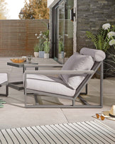 Modern outdoor furniture set including a grey metal frame lounge chair with white cushions, accompanied by a matching grey metal frame coffee table with a round top. The set is displayed on an outdoor patio setting with decorative plants and accessories.