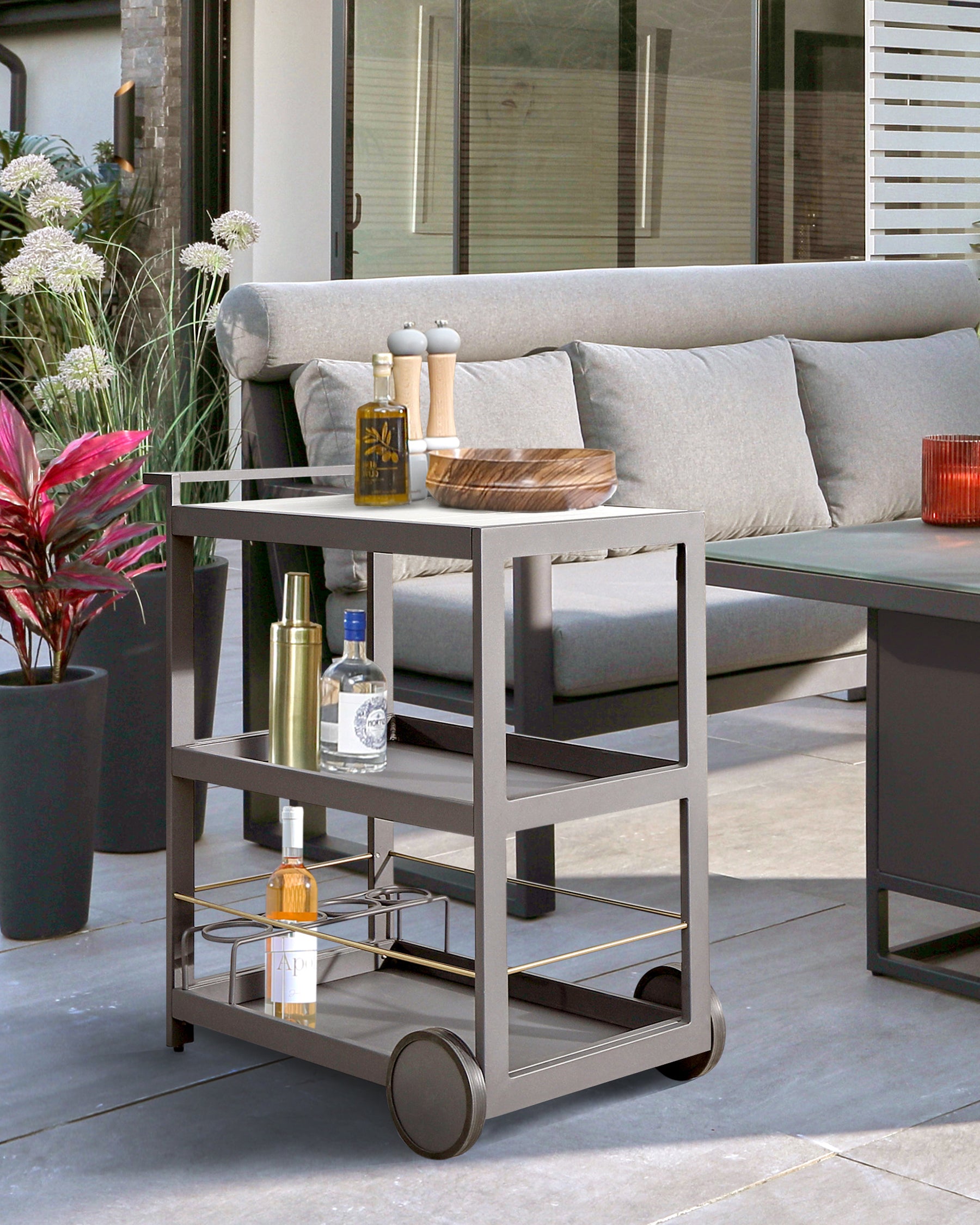 Modern outdoor furniture consisting of a grey metal-framed rolling cart with three shelves and a sofa with a deep grey aluminium frame and light grey cushions. The cart is accessorized with bottles and a wooden tray.