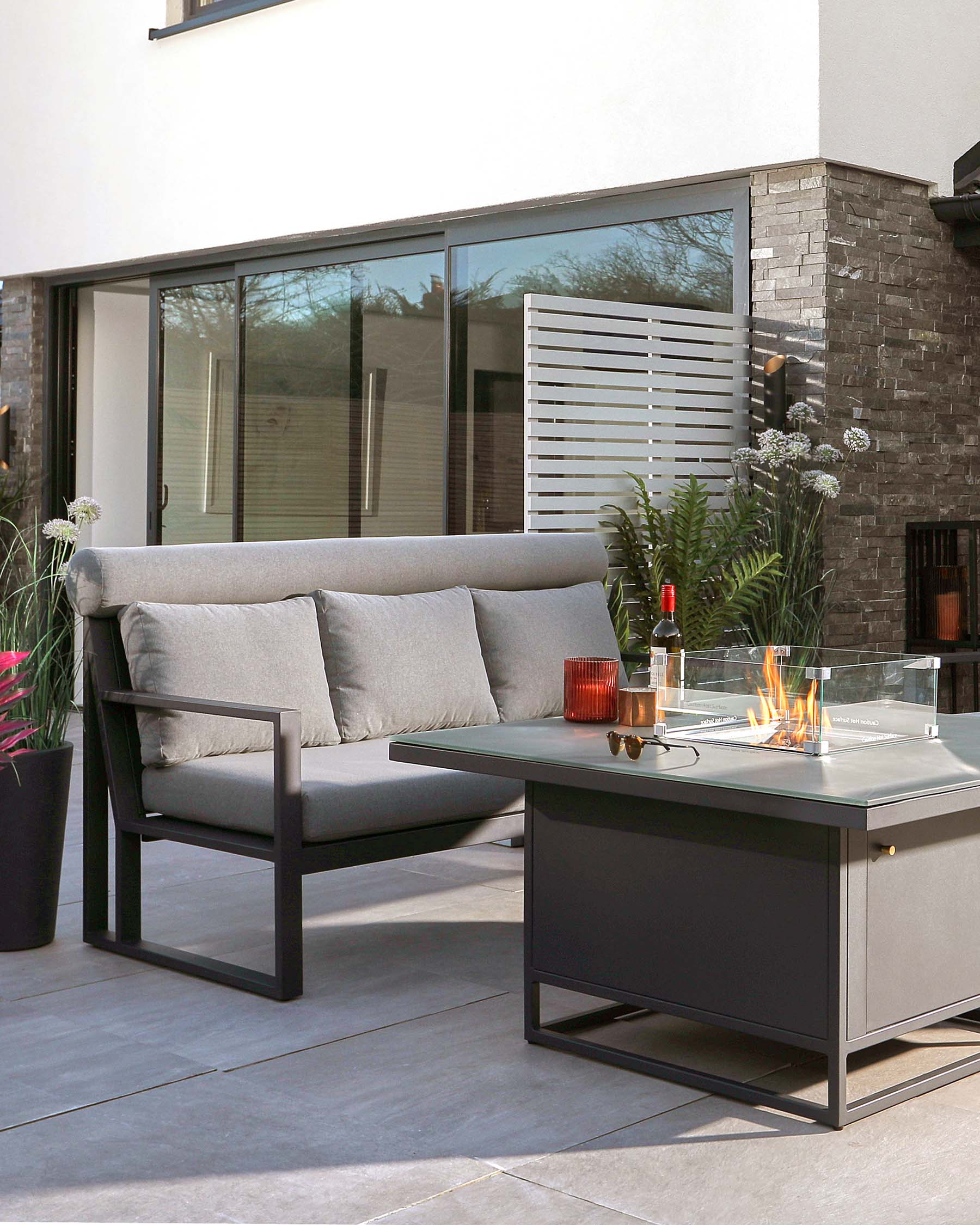 Modern outdoor furniture set featuring a dark grey metal frame loveseat with light grey cushions and a coordinating metal fire table with a glass wind guard and flame in the centre. The setup is complemented by a small black metal side table with a glass top.