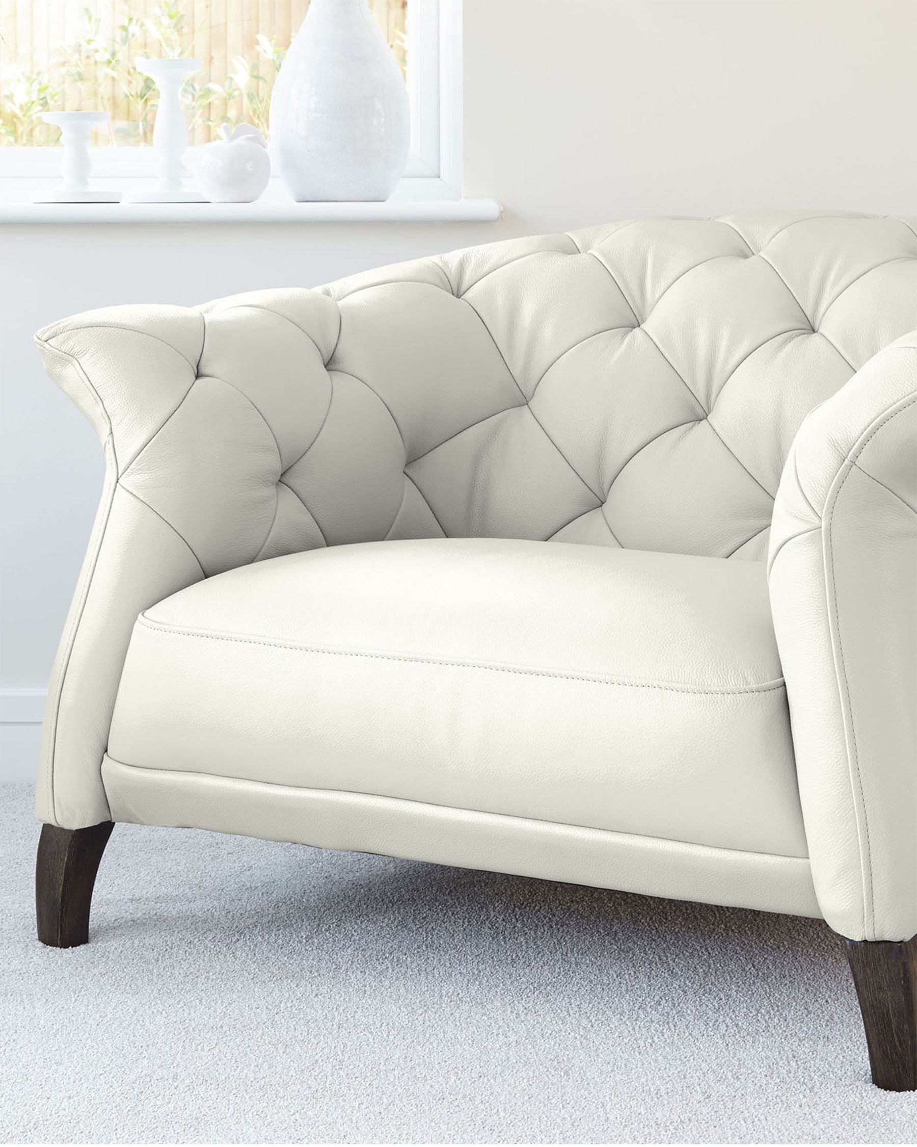 Elegant ivory tufted loveseat with a classic Chesterfield back, plush upholstery, and dark wooden legs on a grey carpeted floor.