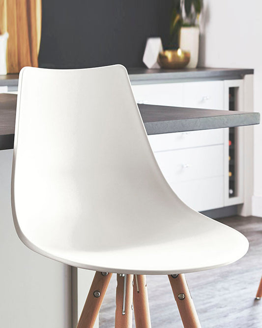 Modern minimalist white chair with a moulded seat and wooden legs supported by black metal rods. In the background, there's a sleek kitchen counter with monochrome cabinetry, accentuated by a decorative vase and geometric figure.