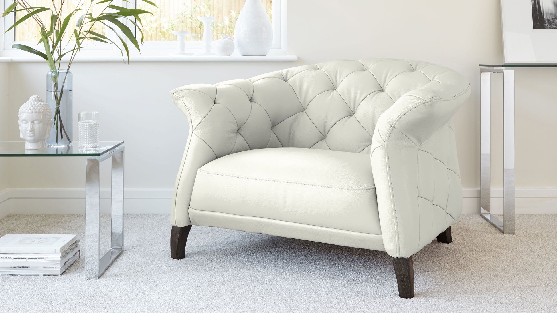 Elegant cream-colored tufted armchair with a high back, overstuffed seating, and dark wooden legs, next to a modern glass side table with a reflective silver base.