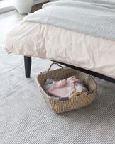 A modern, low-profile platform bed with a dark wooden frame and a chevron-patterned, monochrome throw blanket. Under the bed, there is an artisanal woven basket with two handles, containing soft textiles. The bed and basket are situated on a textured, neutral-coloured area rug.