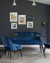 A contemporary dining set featuring a clear glass table with angled black legs tipped with gold accents, and two plush velvet chairs in blue with black legs, also tipped with gold. In the background is a long blue velvet bench with a tufted design, positioned against a dark accent wall.