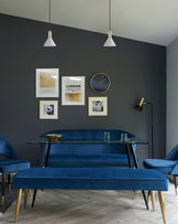 Elegant dining area featuring a rectangular glass-top table with a unique brass-coloured bowl centrepiece, surrounded by luxurious deep blue velvet upholstered seating, including a bench, chairs with curved backs, and gold and black metal accents on the chair and bench legs. The furniture contrasts strikingly against the dark grey wall backdrop.