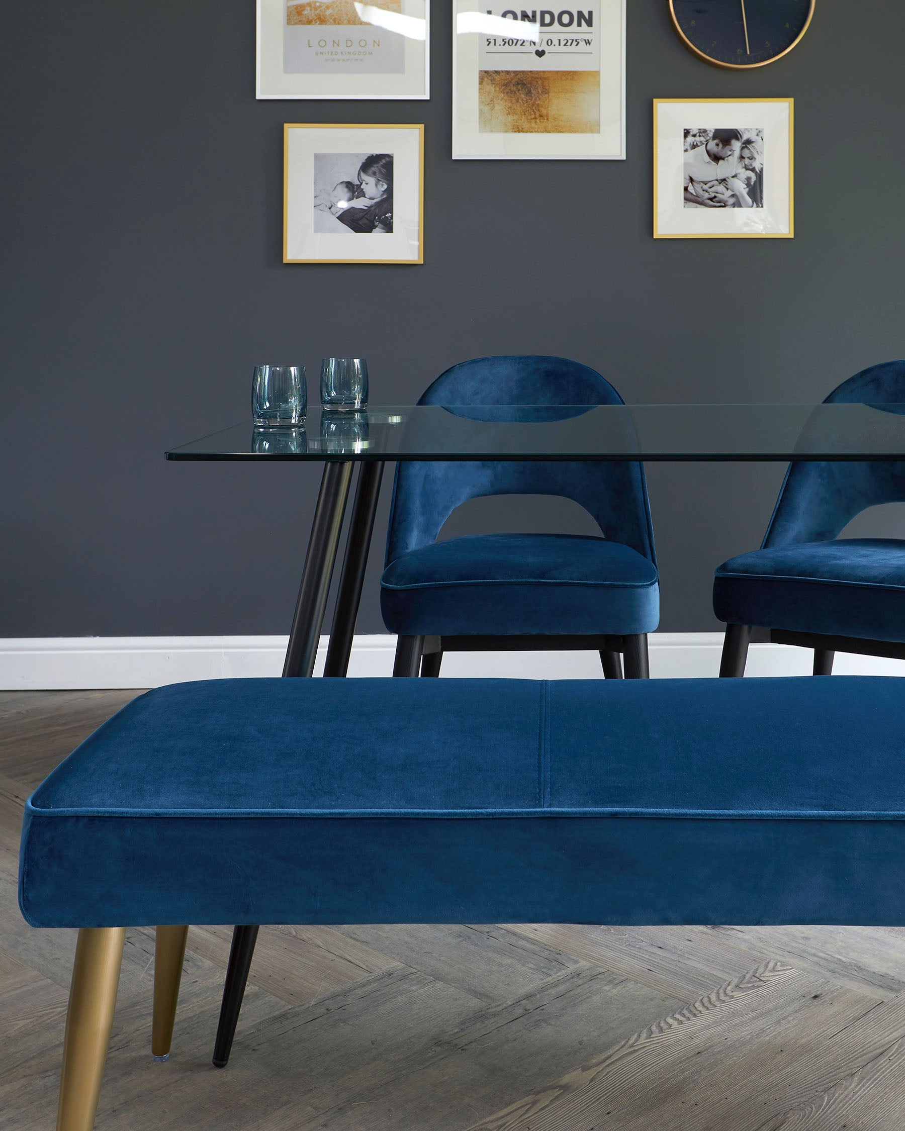 Elegant dining room set featuring a rectangular glass-top table with black metal legs, accompanied by plush velvet blue chairs with a sleek modern design and brass-toned metal legs. In the foreground, a matching blue velvet bench with contrasting golden legs completes the sophisticated ensemble.