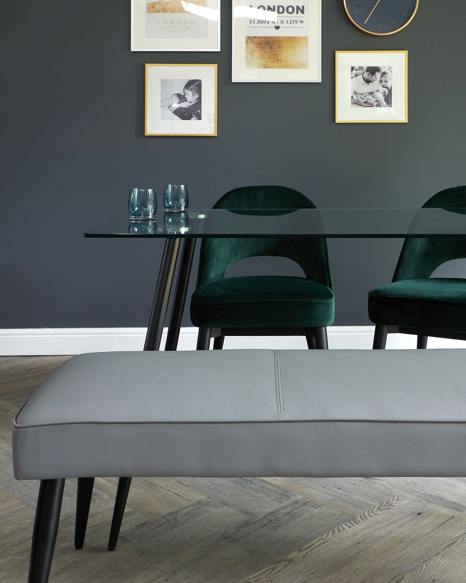 A modern dining room set featuring a sleek, rectangular glass table with black metal legs, paired with sophisticated green velvet upholstered chairs with round backs and matching black legs. In the foreground, a minimalist grey upholstered bench with a subtle stitched detailing and black metal legs compliments the ensemble.