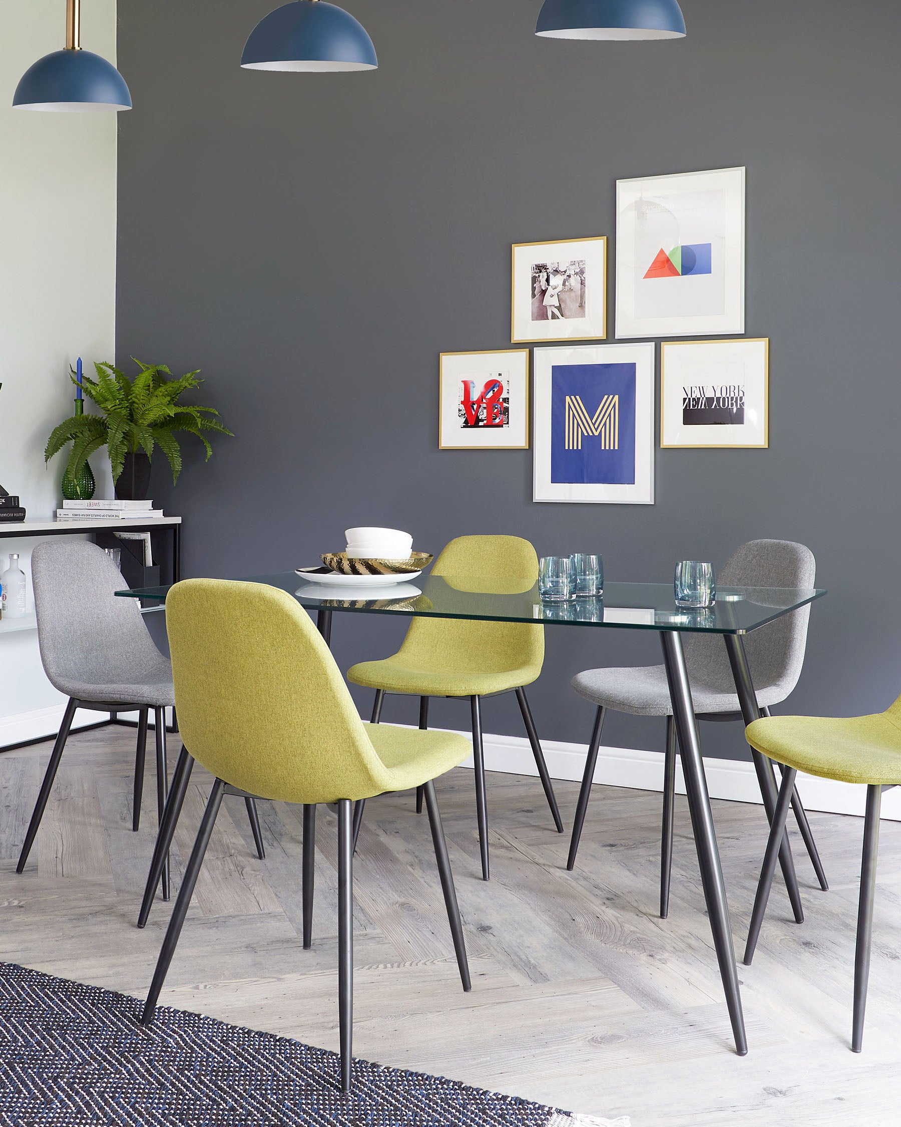 Modern dining room featuring a round glass table with a black metal frame, flanked by four chairs with black metal legs; two chairs upholstered in olive green fabric and two in light grey fabric. A navy blue and white striped area rug lies beneath the set.