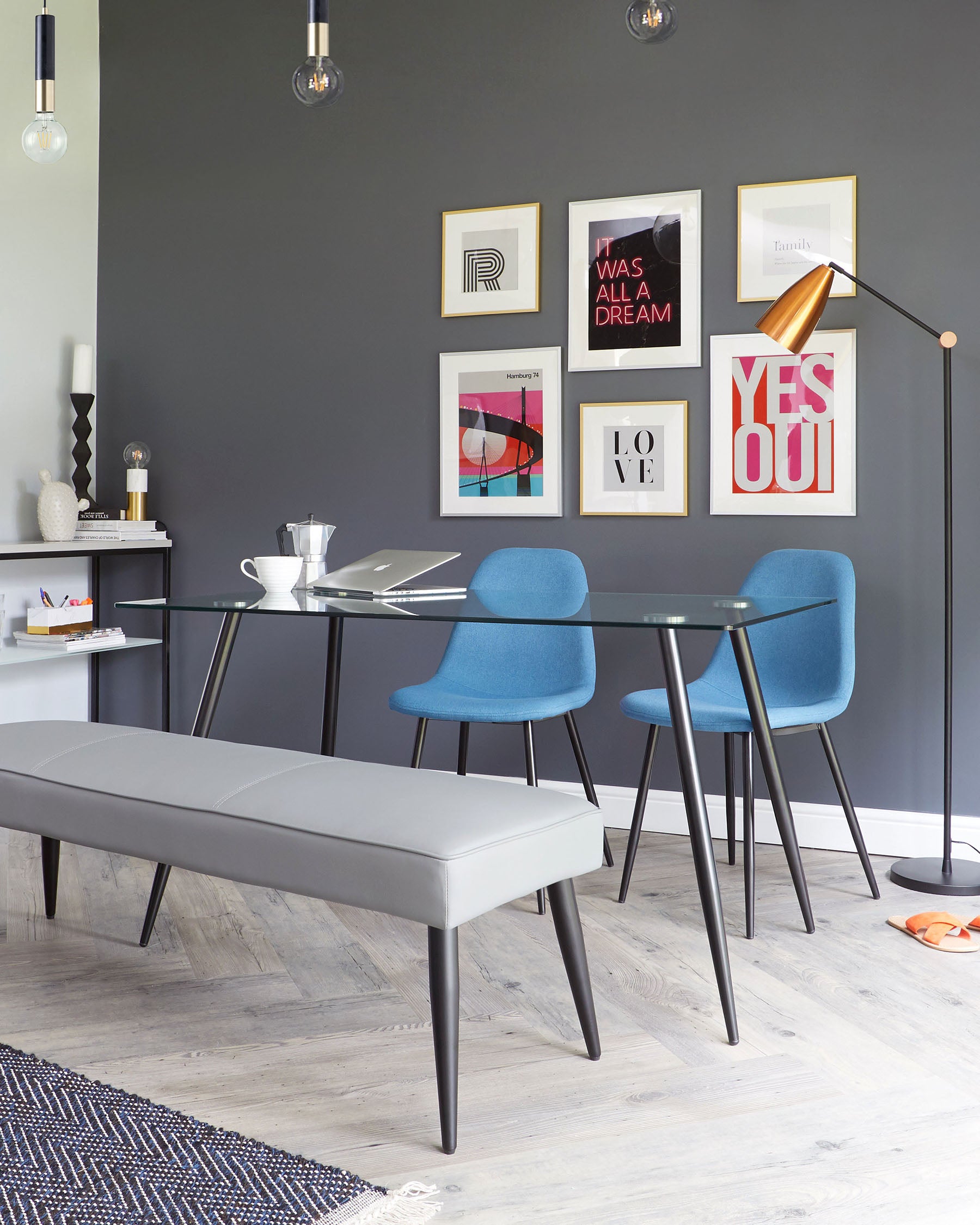 Modern furniture featuring a sleek grey upholstered bench and a pair of vibrant blue dining chairs with black metal legs, flanking a clear glass-top table with a black frame.