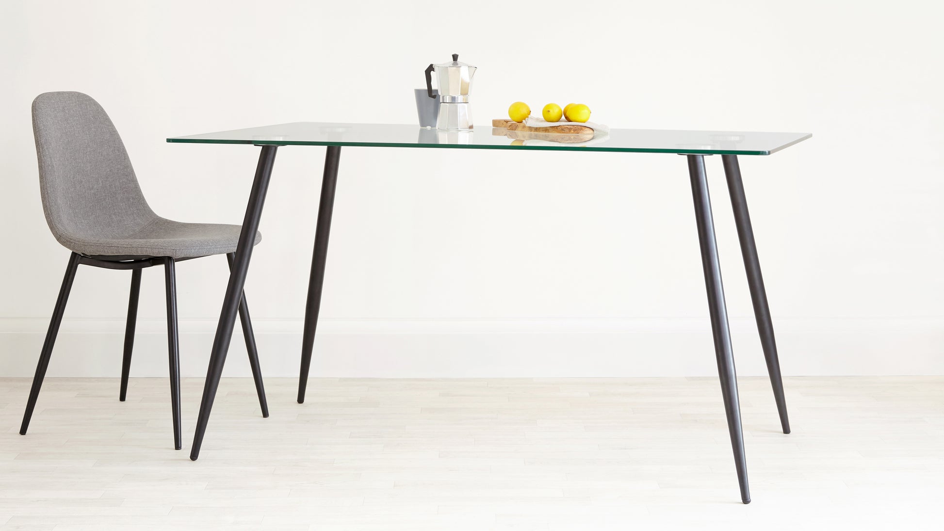 A modern minimalist dining set featuring a rectangular clear glass tabletop with angled black metal legs and a coordinating single grey fabric upholstered chair with slender black metal legs.