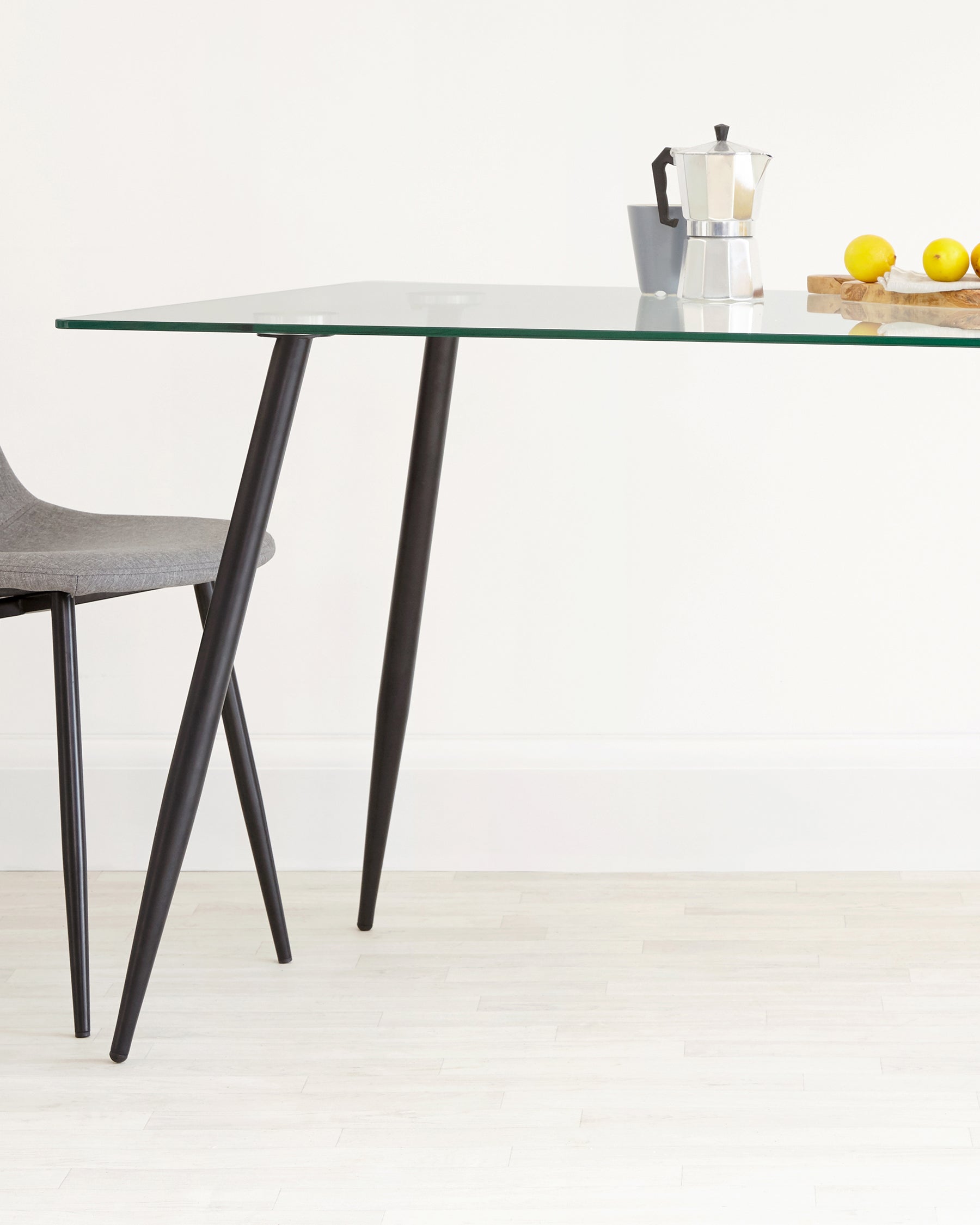 A modern minimalist dining table with a clear glass top and angled black metal legs, accompanied by a grey fabric upholstered chair with black metal legs.