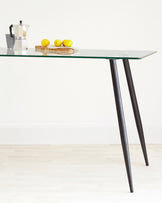 Modern minimalist glass-top table with angled dark legs in a bright interior setting.