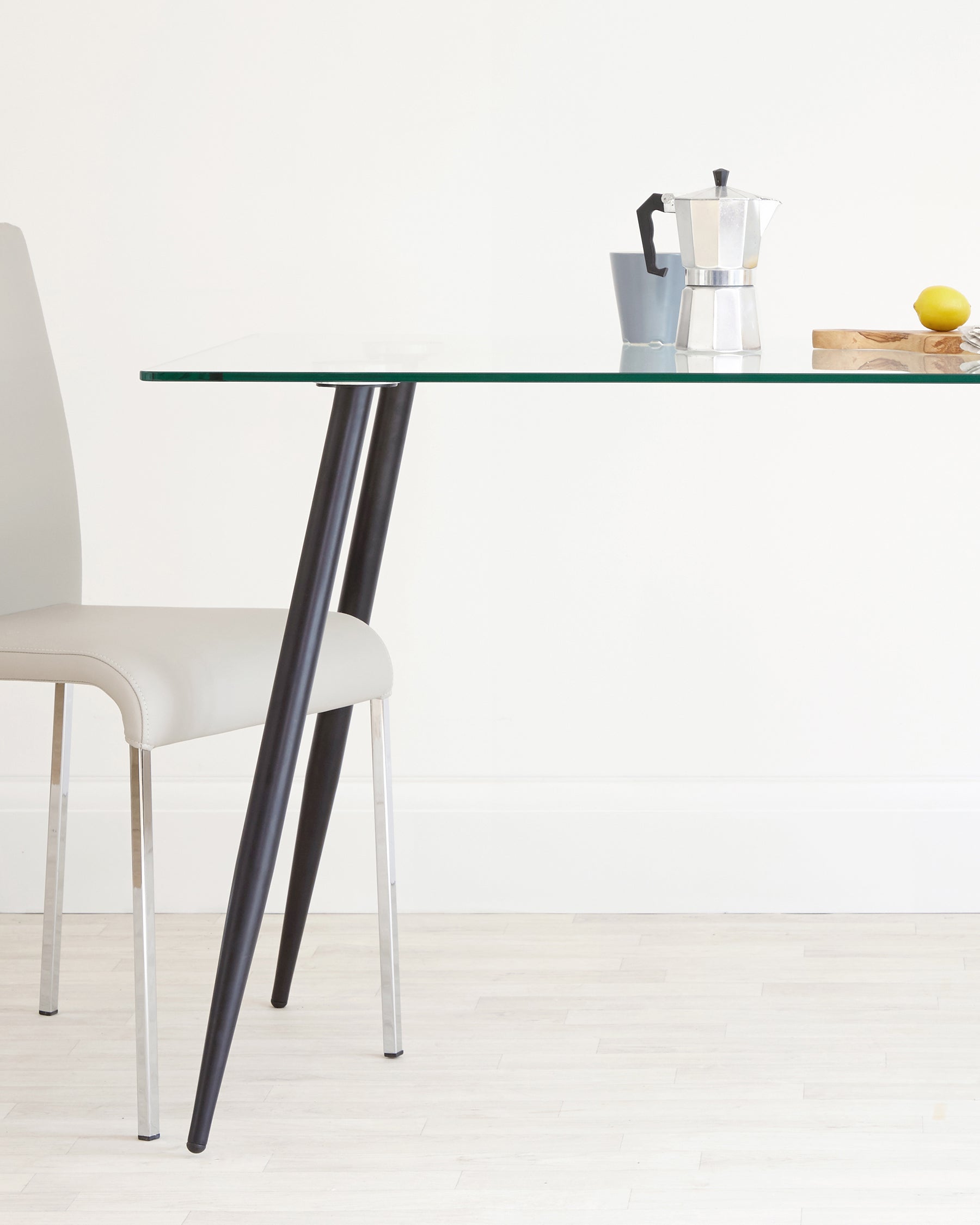 A modern dining setup featuring a clear glass table with angled black legs and a light beige upholstered chair with shiny metal legs.