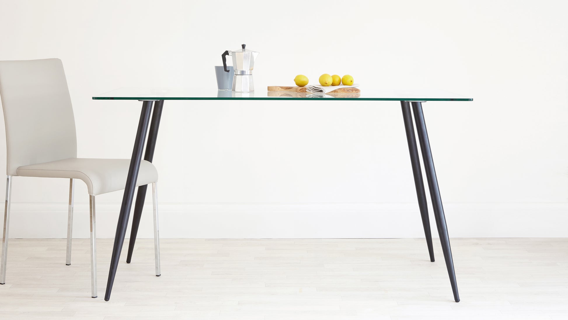 A modern minimalist dining set consisting of a clear glass rectangular table with angled black metal legs, paired with a single cream upholstered dining chair with sleek chrome legs.