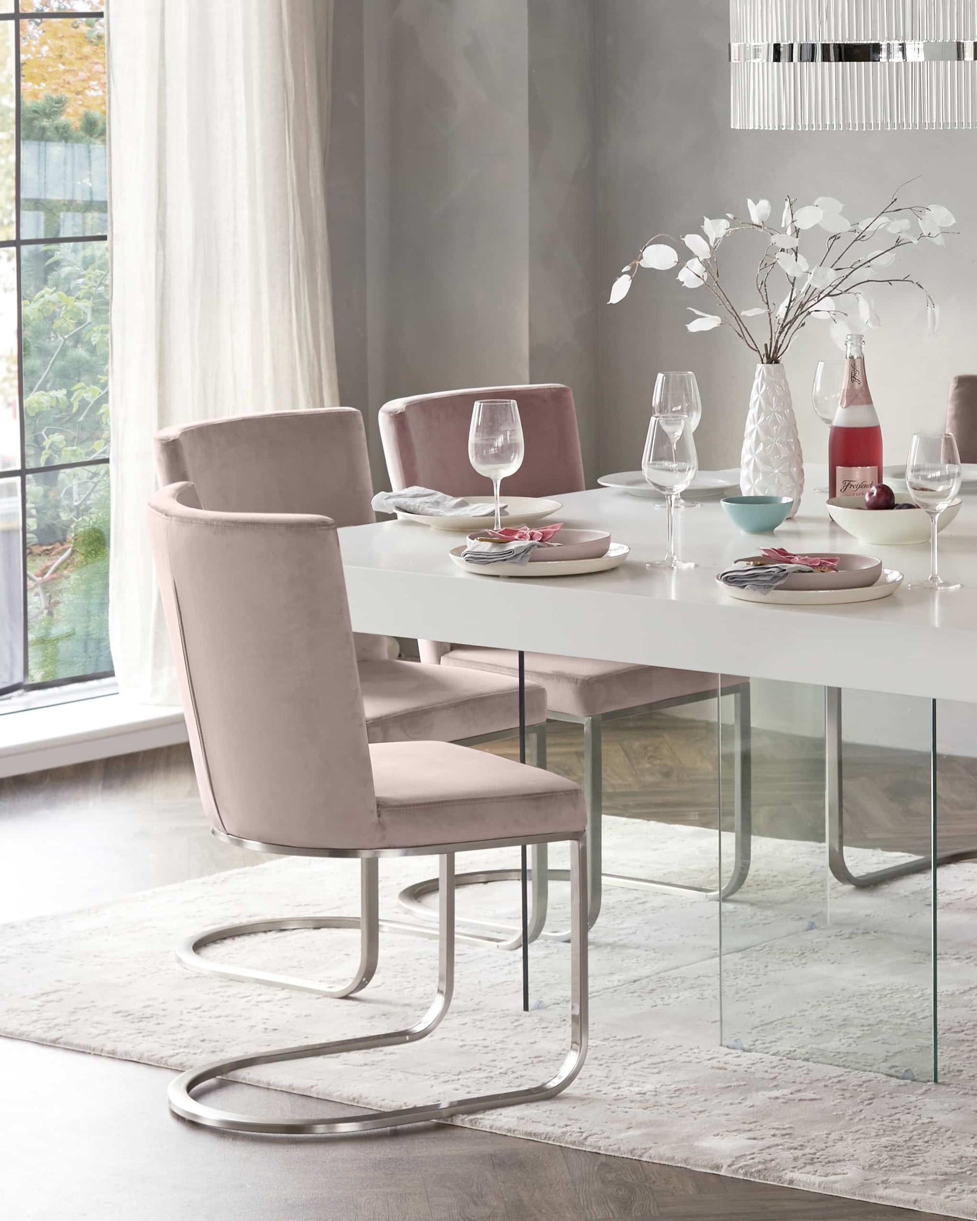 Modern dining room furniture featuring a sleek, rectangular glass-top table with a minimalist white base. The table is surrounded by elegant blush-pink upholstered dining chairs with a smooth, curved backrest and comfortable seating, all supported by stylish metallic silver cantilevered frames. The setting conveys a contemporary yet inviting atmosphere.