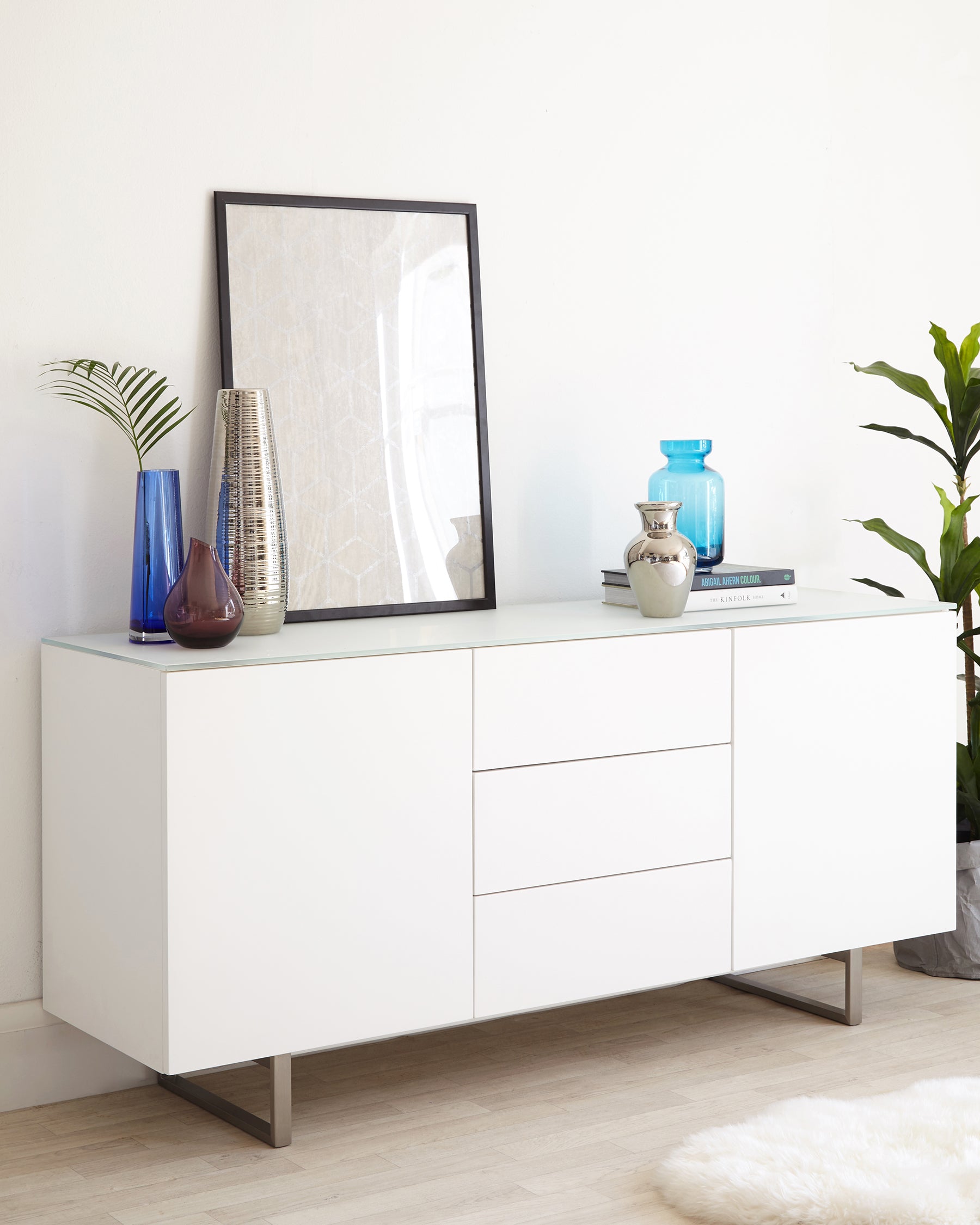 Modern white sideboard with sleek lines and metal leg accents. Features two cabinet doors and three drawers for storage, with a glossy finish. The top surface is decorated with various vases and a framed artwork.