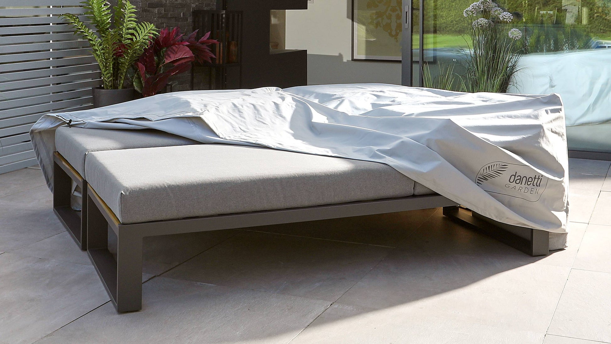 A modern outdoor sun lounger with a sleek, low-profile metal frame in a dark finish and a light grey upholstered cushion. It includes a partially rolled white cover with the logo "Danetti Garden" on the front. The lounger is set on a patio with plants in the background, suggesting an inviting outdoor environment.