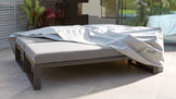 Modern outdoor double sunbed with a grey cushioned mattress on a sleek, dark frame, partially covered with a white protective cover featuring the logo 'Danetti Garden'. The sunbed is set on a patio area with greenery in the background.